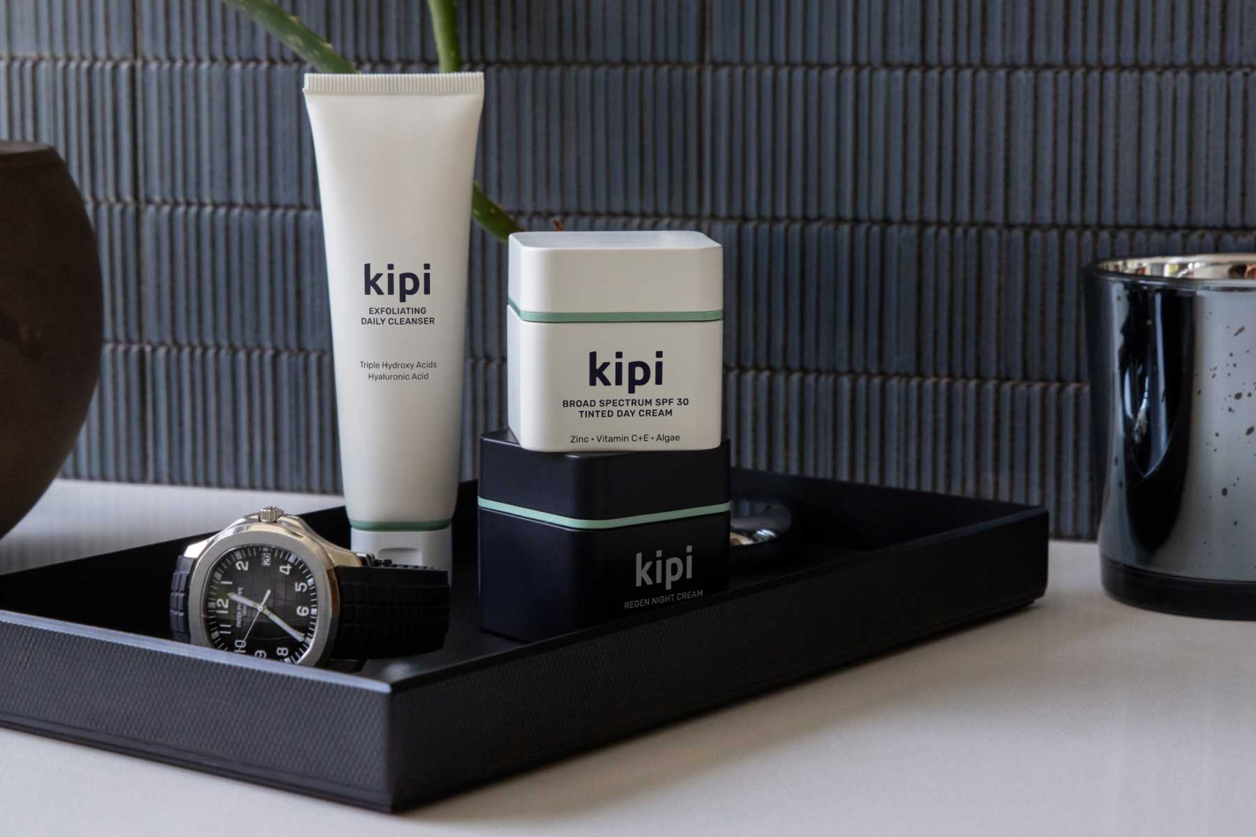 Kipi skincare products on a black tray with a watch and plant against a dark tiled wall.
