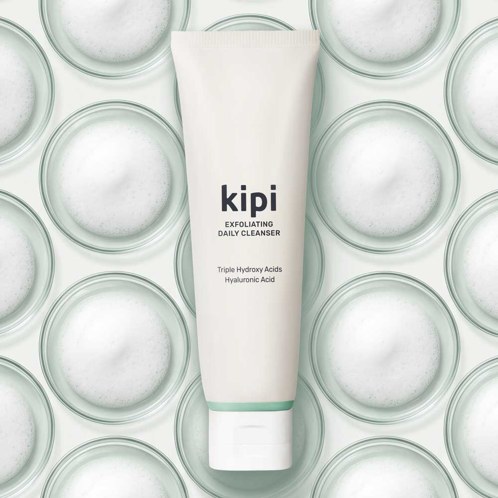 A tube of Kipi Exfoliating Daily Cleanser floating above a background of small glass dishes full of foam from lathered face cleanser.