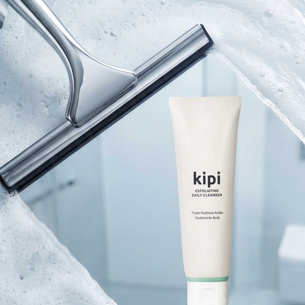 A squeegee sliding across a glass shower door that is covered in suds, revealing Kipi Exfoiating Daily Cleanser through the clear glass.