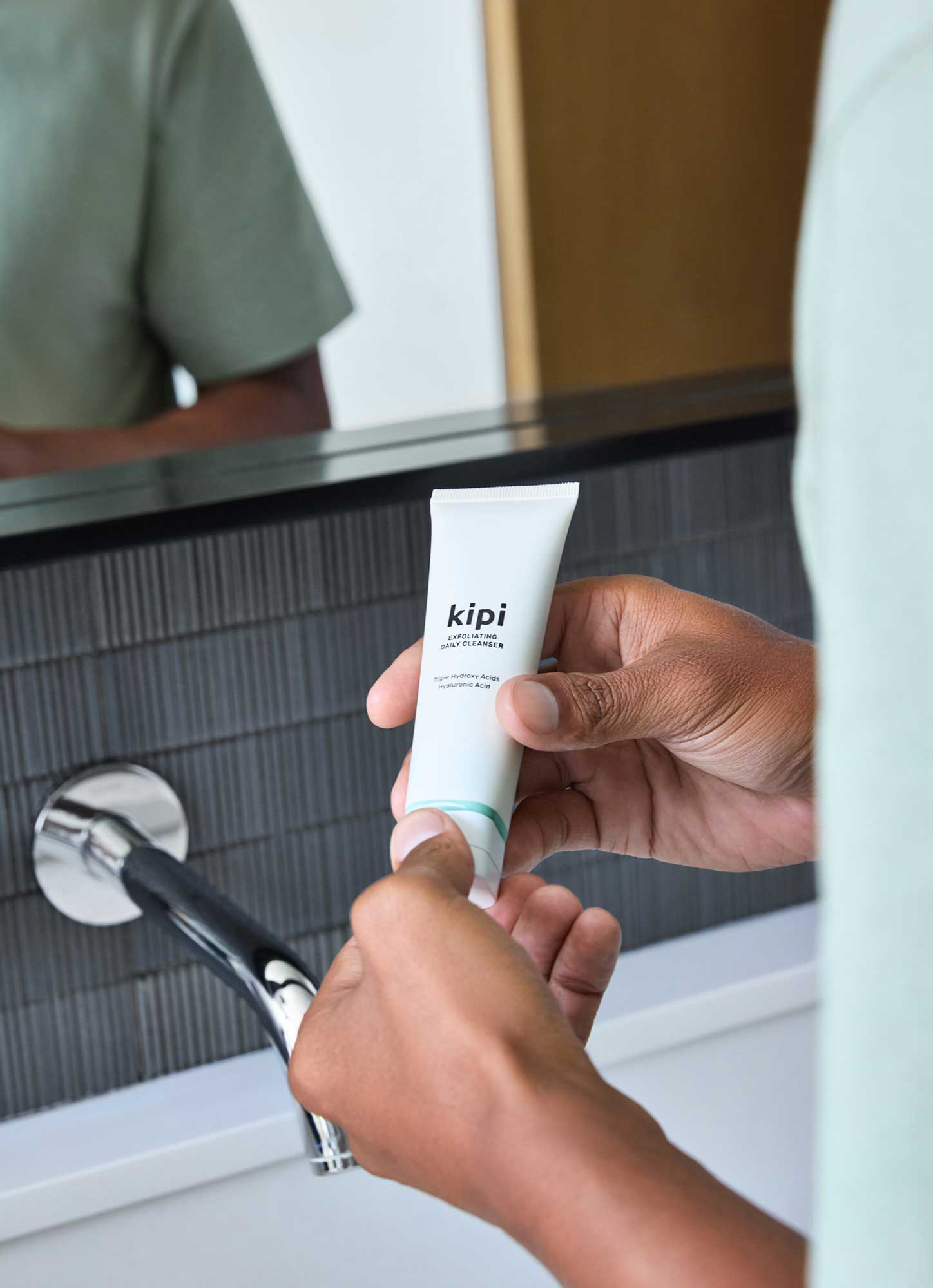 Person holding a tube of Kipi Exfoliating Daily Cleanser in front of a mirror.
