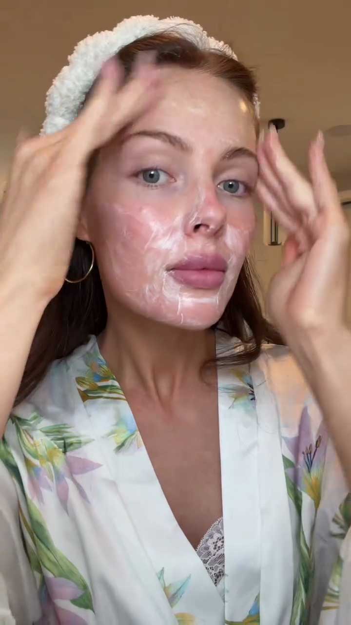 Woman applying a facial cleanser with a headband on her head, wearing a floral robe.