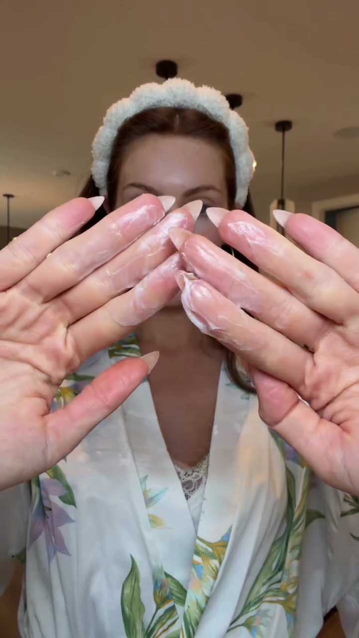 Person wearing a floral robe with hands covering the face, showing off manicured nails and cleasner lathered on her hands.